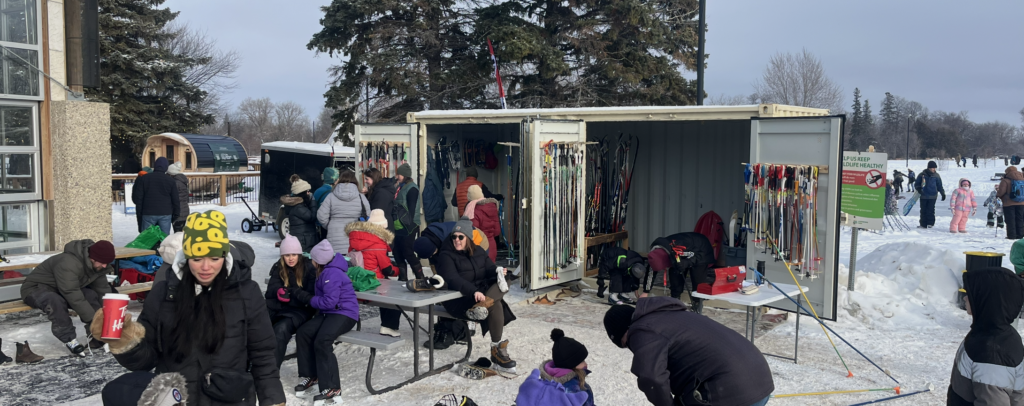 a beige shipping container with side doors that are open to show lots of ski equipment inside and a large crowd of people in front and snow and evergreen trees in the background